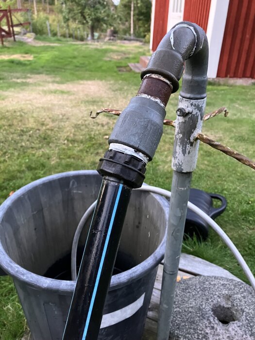 Bilden visar en koppling med läckage mellan ett svart rör och ett järnrör vid en utomhusinstallation, nära en stor hink.