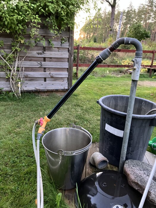 Borrad brunn med svart rör och slang kopplade till hink och tunna i trädgård, med synliga läckande kopplingar i kvällsljus.