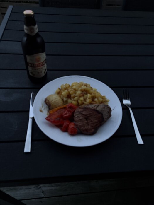 Grillad oxfilé med grillade grönsaker, råstekt potatis och en flaska Mariestads öl på ett bord utomhus i skymning.