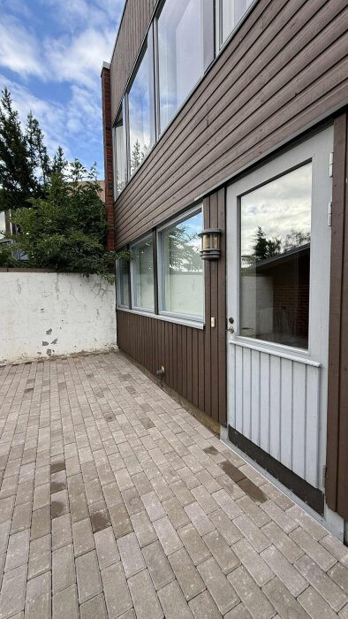 Brun träfasad med fönster och en ytterdörr. På fasaden hänger en modern lampa. Stenlagd uteplats och vit murad vägg bredvid.