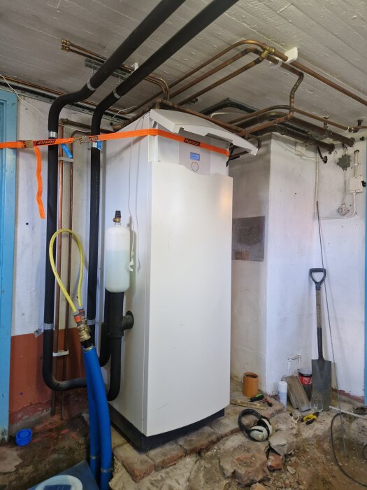 Vitt skåp i källare med rör och spännband fastsatt runt. Verktyg och slangar synliga på golvet bredvid.