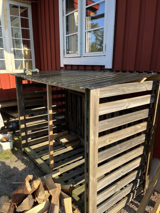 Vedskjul byggt av träplankor mot en röd husvägg, med staplad ved på marken framför.
