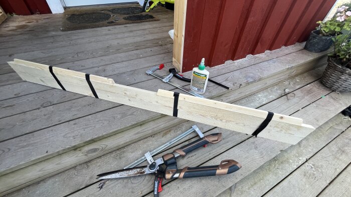 Träbräda med svarta remmar, verktyg och trälim på altan framför rött hus.