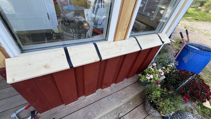 Träfönsterbleck i konstruktion med eltejp på ett rött hus; blommor och en blå tunna syns framför fönstret.