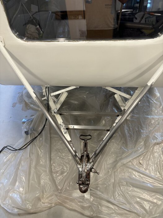 Vitt släp eller husvagn med dragkrok och stag över en plastskyddad golvyta i ett garage eller verkstad.