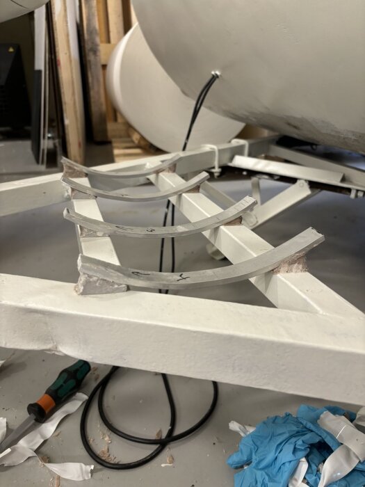 En vit träställning med böjda metallstänger i ett garage, omgiven av verktyg och blå arbetsvantar på golvet.