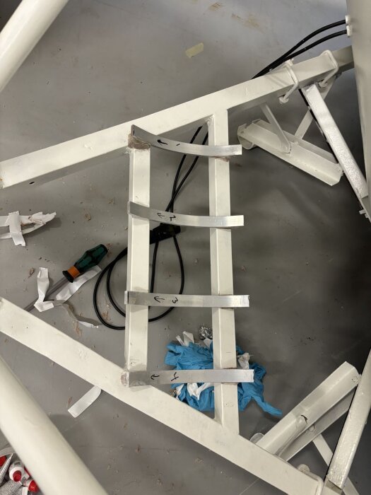 Vitt metallställ med pilar och markeringar, omgiven av verktyg och skräp på golvet.