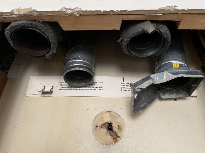 Tre exponerade ventilationsrör utan köksfläkt, där isoleringen är i slitage under en köksö, bredvid en etiketterad bruksanvisning.