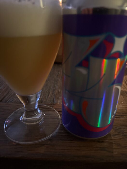 Ett glas öl bredvid en färgglad Omnipollo-burk på ett träbord.