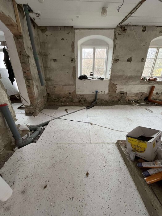 Ett gammalt stenhus under renovering med avskalade väggar, rörinstallationer och verktyg på fönsterbrädan; golvet av vita skivor; murbruk saknas.