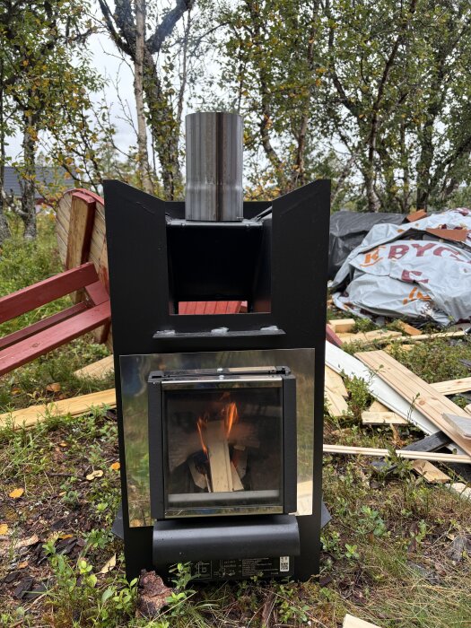 Svart bastukamin med synlig låga, omgiven av byggmaterial och träd i bakgrunden i en utomhusmiljö.