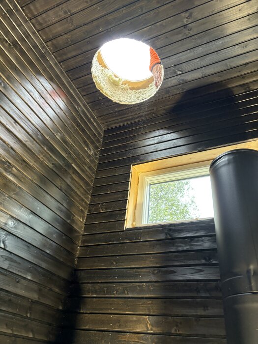 Öppning för skorsten i taket på en nybyggd bastu med mörka träväggar, ett fönster och en del av kaminen synlig.