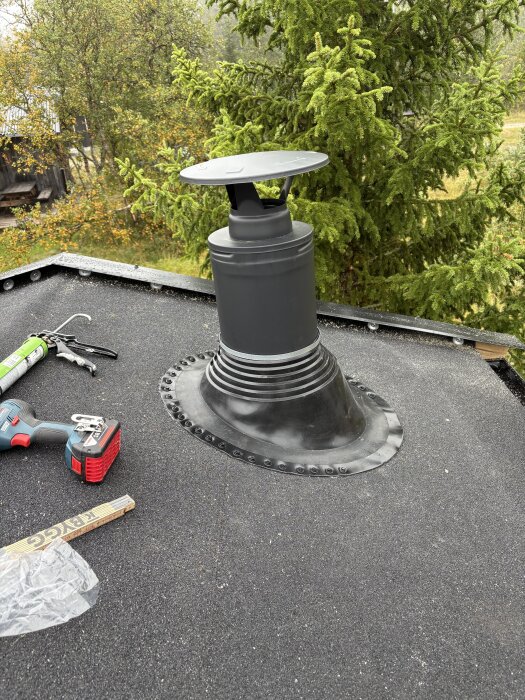 Svart skorsten installerad på en grå takyta, med verktyg och material runtomkring. Skog i bakgrunden.