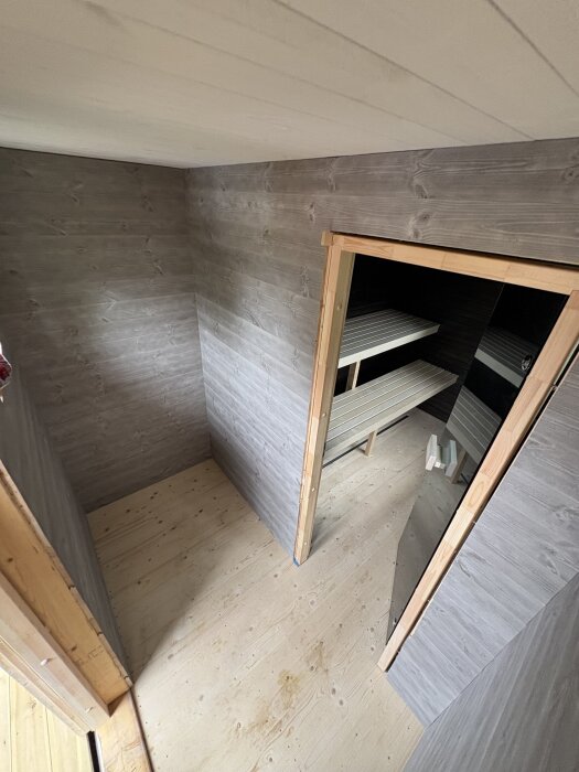 Bild av ett halvfärdigt basturum med träpanel och lavaleder, sett från ovan, med ljus träskiva på golvet och öppning till bastudörr.