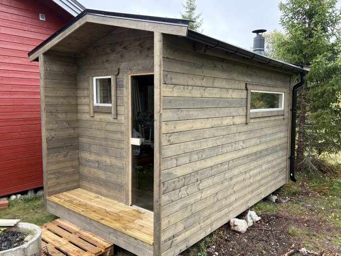 En nybyggd bastubyggnad med träpanel och skorsten, delvis omgiven av träd. Ytterpanelen är i neutral träfärg och förrummet saknar dörr.