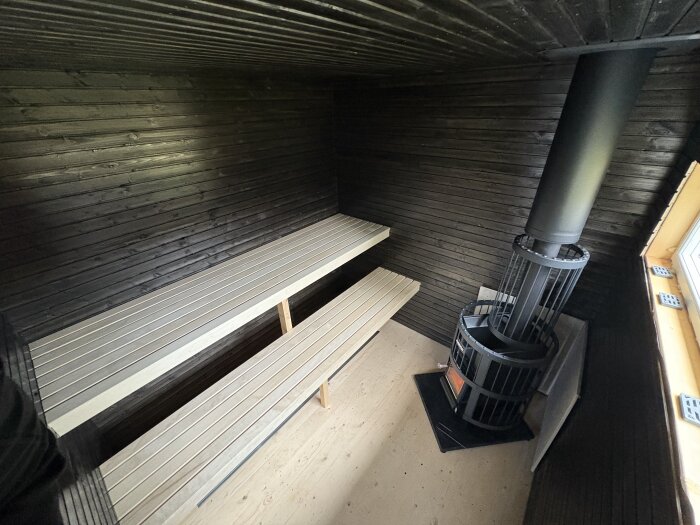 Bastu med monterad kamin och bänkar mot en mörk träpanel. Kamin klart för inspektion, bänkar stödja av ett extra ben för stabilitet.