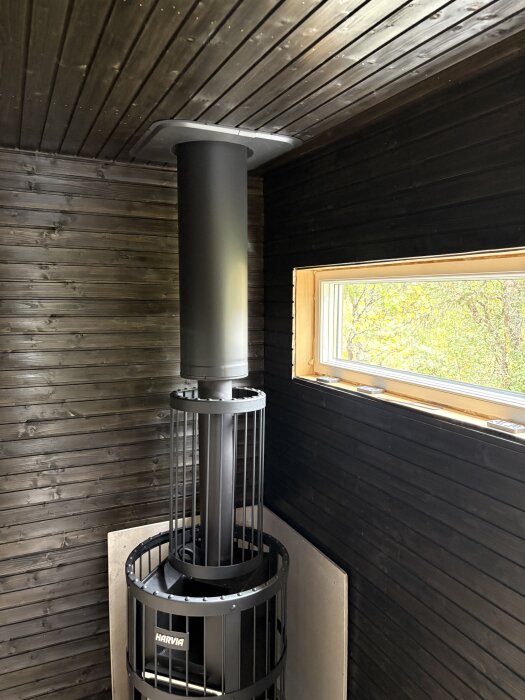 Svart bastukamin med skorsten i ett rum med mörka träväggar och ett fönster som visar grönska utanför.