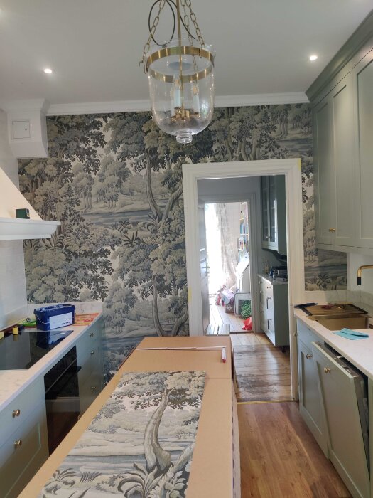 Kök med tapetserade väggar i naturmotiv, grönaktiga köksskåp och ett hängande lamparmatur i glas och mässing.