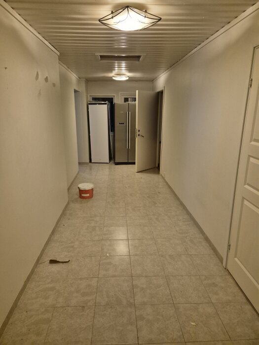 Långsmal hall med grått klinkergolv, takfönster, taklampor och vitmålade väggar. En vit hink och ett verktyg på golvet.