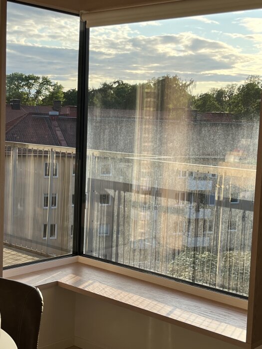 Fönster med svår borttagen beläggning syns tydligt mot dagsljus; utsikt över flera byggnader och träd i bakgrunden.