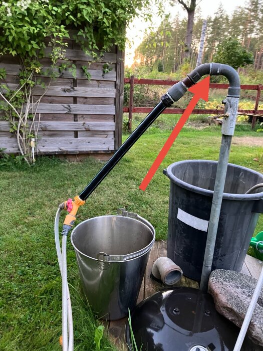 Utomhusinstallation med metall- och plastledningar, en hink och slangkoppling. En röd pil pekar mot en böjd del av röret.