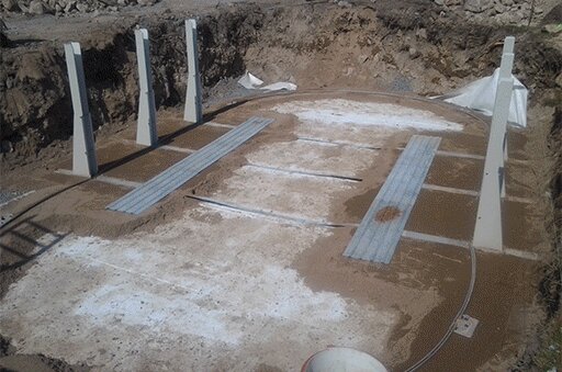 Grävd grop med påbörjad konstruktion av en pool eller terrass, med betongstöd och jordiga omgivningar.