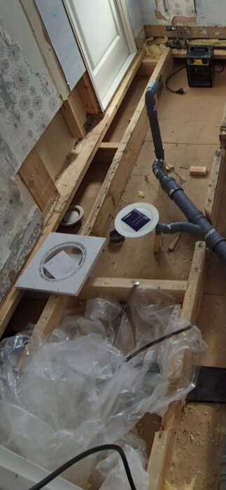 Renovering med öppet trägolv, rörinstallationer, och plastark. Papper och verktyg ligger utspritt. Kakelarbete pågår.