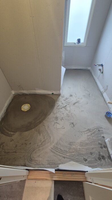 Ett rum med grå betonggolv, väggar av gipsskivor och ett golvavlopp under pågående renovering, fotspår synliga i cementen.