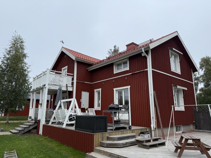 Nymålat rött trähus med vit uteplats, gungställning och grill på trädäck. Klart innan regn; vitt och garage återstår.