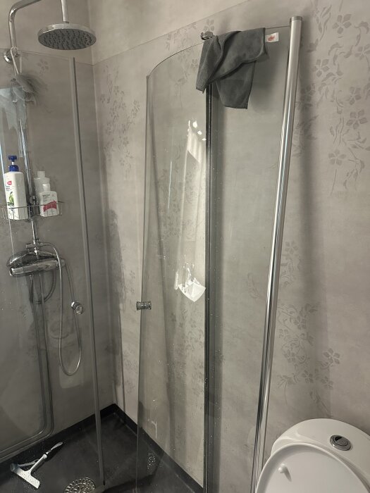 Duschhörna med glasvägg och duschset, trasiga delar syns inte tydligt, handduk hänger över kanten, badrumsdetaljer i bakgrunden.