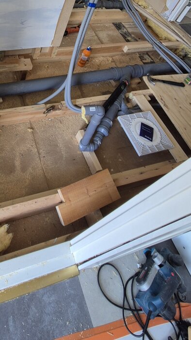 Byggarbetsplats med synliga rör, verktyg och träreglar, del av golvkonstruktion, fläkt och isolering i bakgrunden.