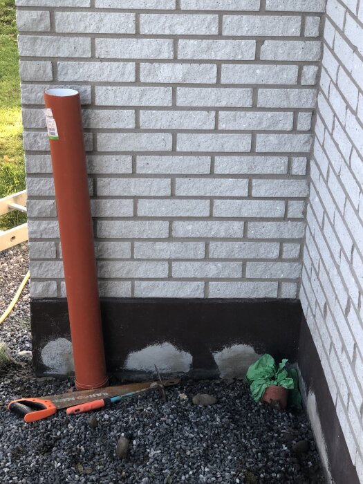 Brun metallrör och såg vid hörn av tegelvägg med grusig mark. Grön plastpåse vid väggen, nytt murverk synligt vid botten.