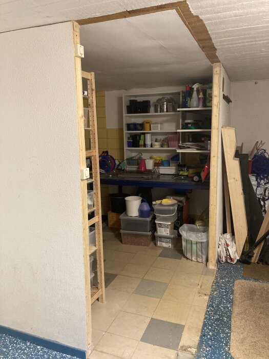 Öppen snickarbod med hyllor fyllda med förvaringslådor och verktyg. En del av väggen har rivits för access.