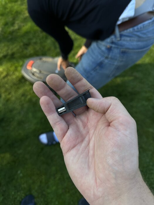 Hand håller en svart plastdel med en liten metallbit, bakgrund visar en person böjd över en robotgräsklippare på en gräsmatta.