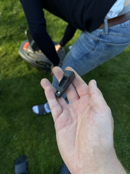En hand som håller en liten svart del med metallkomponent, personen står bredvid en robotgräsklippare på grönt gräs.
