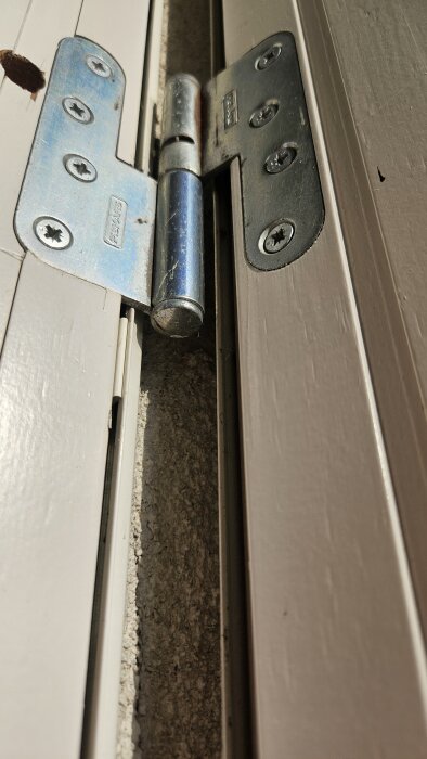 Närbild av ett dörrgångjärn där skruvar i karmar ser slitna ut och hålen är rundade. Gångjärnet saknar justerskruvar.