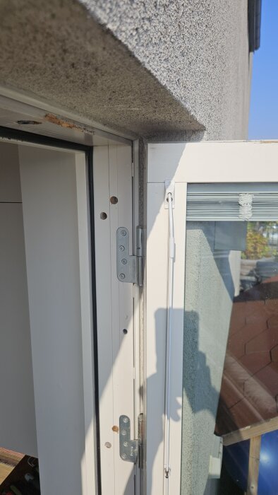 Altandörrens gångjärn och karmsida med runda skruvhål och slitningar, mot en ljus fasad.