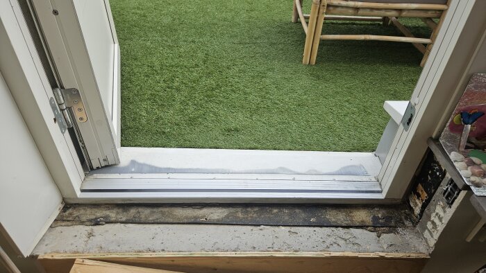 Altandörrens bottenkarm med tecken på fuktskada och svartmögel, omgiven av konstgräs och bambumöbler utomhus.