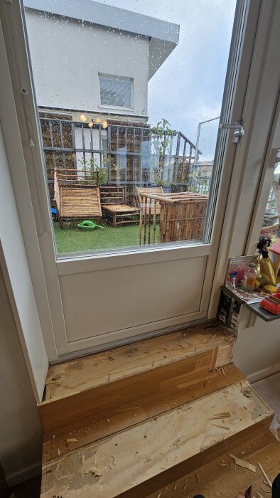 Fuktigt trägolv vid altandörr med spår av parkettskador och regndroppar på fönstret, utsikt mot uteplats med trädäck och växter.