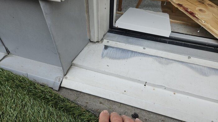 Altandörrens tröskel med synliga fukt- och mögelskador, delvis gömt av en borttagen del av parkettgolvet, synligt gräs nedanför basen.