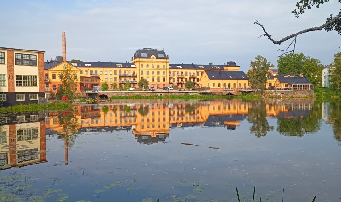 Fabriksbyggnad speglas i en lugn å, med gul fasad och skorsten; nu ombyggd till bostäder med exteriör bevarad.