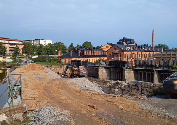 Byggarbetsplats vid Tunafors kraftstation med grävmaskin och damm under renovering, fabriksliknande byggnader i bakgrunden.