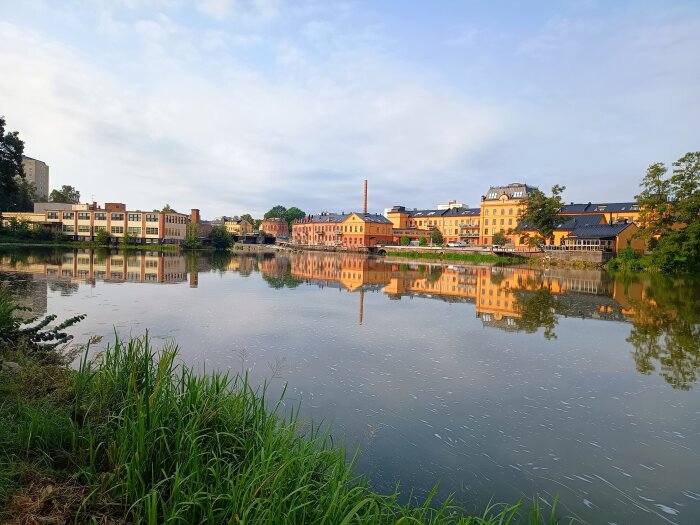 Stadsmiljö vid en vattenspegel med byggnader och grönska som reflekteras i den lugna ytan, skapar en fridfull oas mitt i centrum.