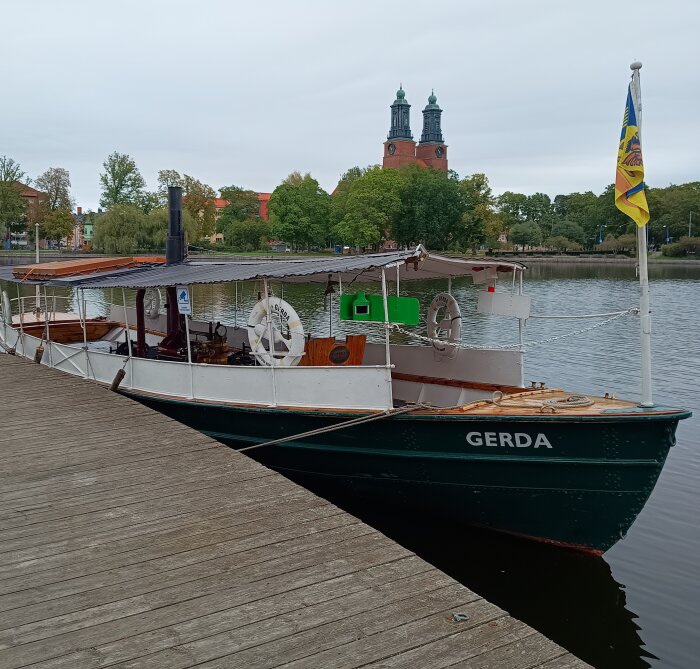 Ångslupen Gerda ligger vid kajen i Eskilstunaån, omgiven av grönska och en kyrka i bakgrunden.