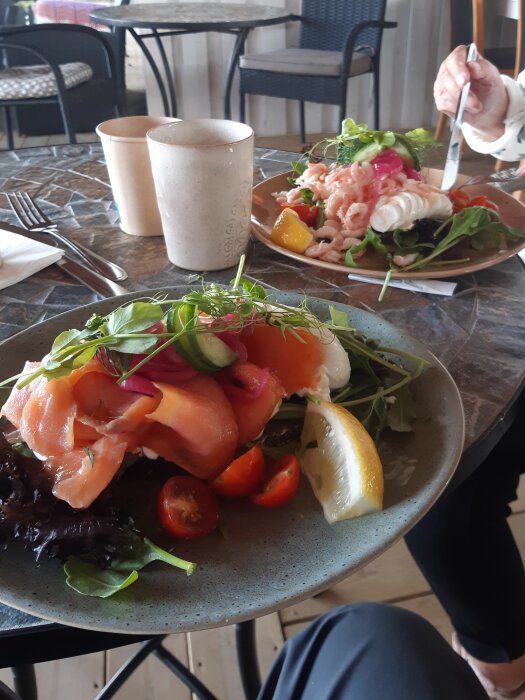 Tallrik med lax, räkor, grönsaker och citronklyfta på ett matbord i en restaurangmiljö.
