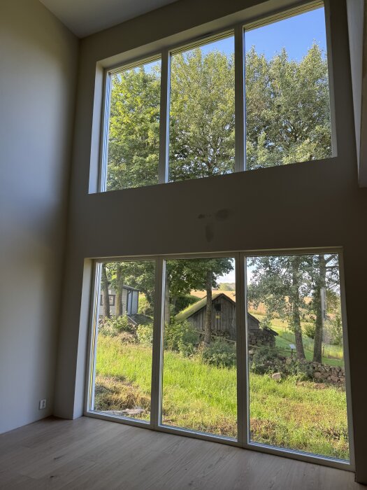 Stora fönsterpartier i ett vardagsrum med utsikt över en grön trädgård och en ladugård. Ljuset strömmar in från de stora fönstren.