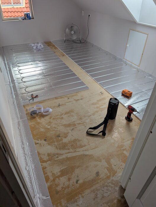 Golvvärmeinstallation med LK EPS 18-skivor i ett rum, inklusive silvertejpade skarvar och verktyg. En fläkt och annan utrustning syns i bakgrunden.