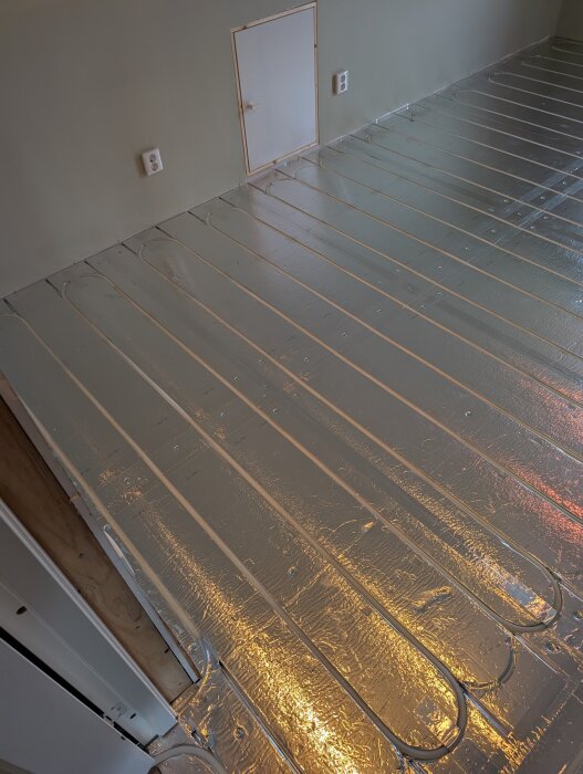 Golvvärmesystem installerat med aluminiumöverdragna EPS-skivor, rör lagda i utrymmen. Vägg med två eluttag och ett vitt skåp syns.