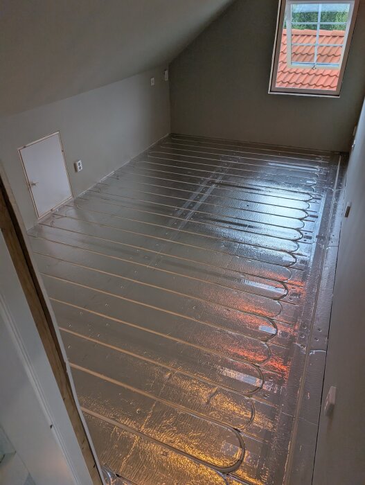 Golvyta täckt med silvriga golvvärme-skivor av typen LK Combiboard EPS 18, med inlagd slang för golvvärme, under ett fönster i ett rum.