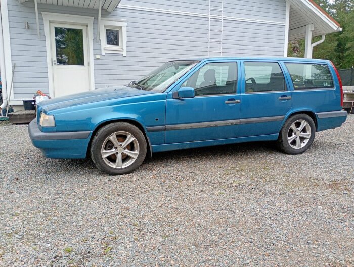 En blå Volvo kombi parkerad på en grusuppfart framför ett grått hus. Bilens rails behöver nya skruvar. En blinkers saknas.